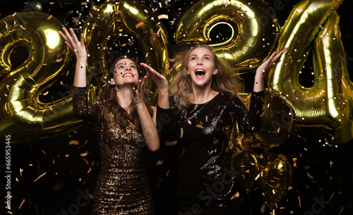 Happy gorgeous girls in stylish sexy party dresses holding gold 2024 balloons, having fun at New Year's Eve Party.