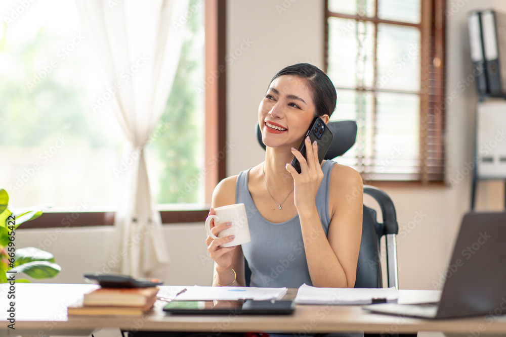 Asian Woman entrepreneur busy with her work in the office. Young freelancer Asian woman talking over smartphone or cellphone while working on computer at her desk. Asian Woman entrepreneur busy with her work in the office. Young freelancer Asian woman talking over smartphone or cellphone while working on computer at her desk.