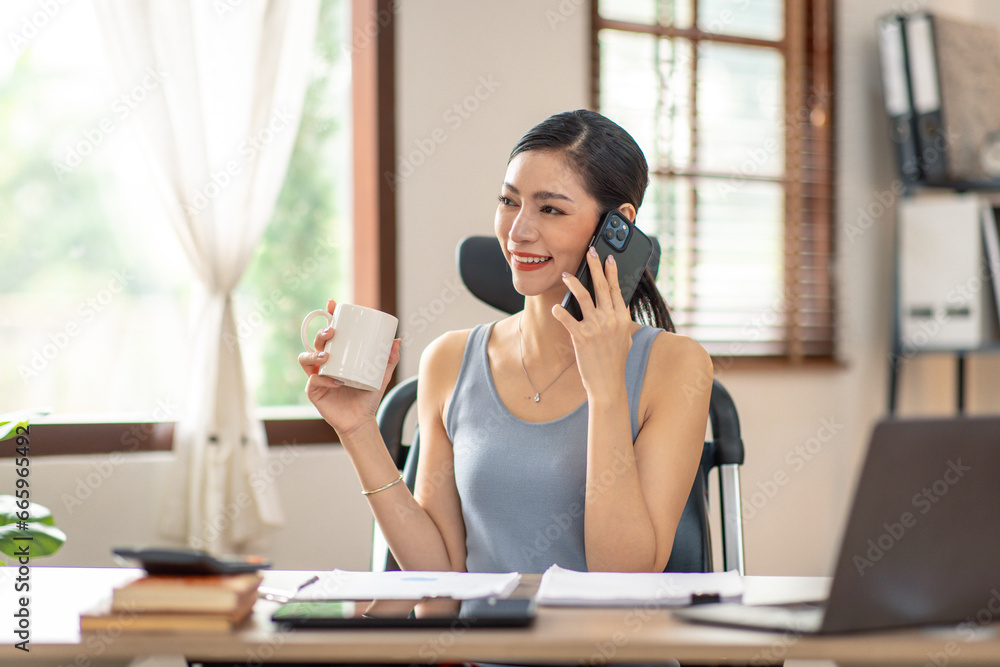 Asian Woman entrepreneur busy with her work in the office. Young freelancer Asian woman talking over smartphone or cellphone while working on computer at her desk.