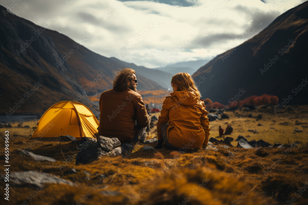 Happy couple in outwear sitting on the rocks near the tent camp against the backdrop of picturesque mountain landscape. Caucasian family is engaged in a mountain hiking. Tourism and active lifestyle.