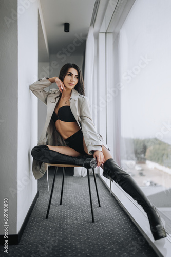 A stylish beautiful young brunette girl with curly hair, an Armenian woman in black underwear and a coat sits in a hotel room indoors on a chair near the window. Photography, portrait.