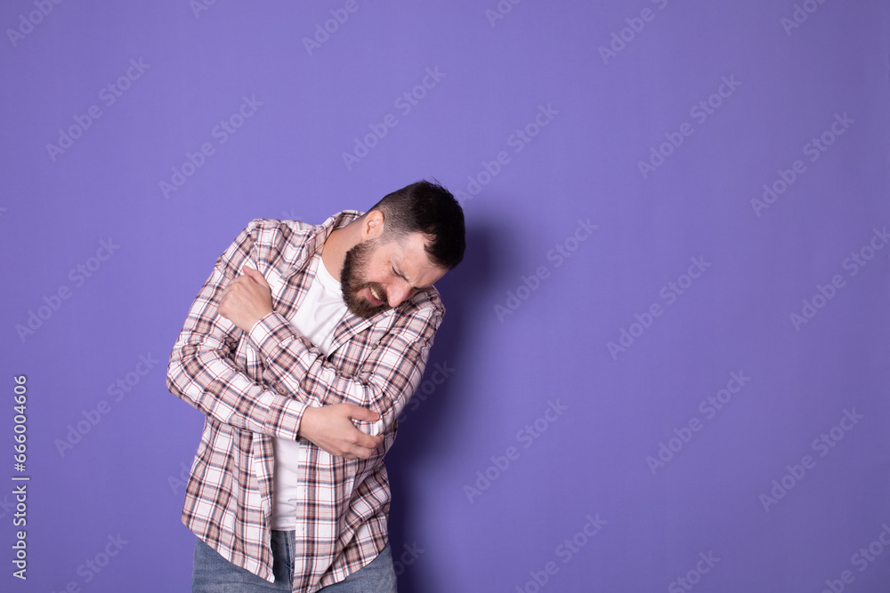 Portrait of bearded man massaging aching elbow, frowning from acute pain, strain and strain problem, trapped nerves, rheumatoid arthritis. Indoor studio shot isolated on purple background. Portrait of bearded man massaging aching elbow, frowning from acute pain, strain and strain problem, trapped nerves, rheumatoid arthritis. Indoor studio shot isolated on purple background.