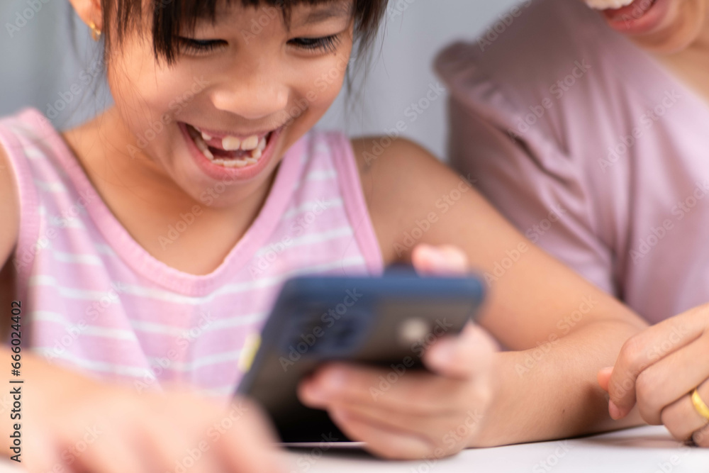 Happy Asian family mother and cute daughter using phone together at home. Mother and little girl laughing happily using smartphone watching funny social media video app sitting at table together. Happy Asian family mother and cute daughter using phone together at home. Mother and little girl laughing happily using smartphone watching funny social media video app sitting at table together.