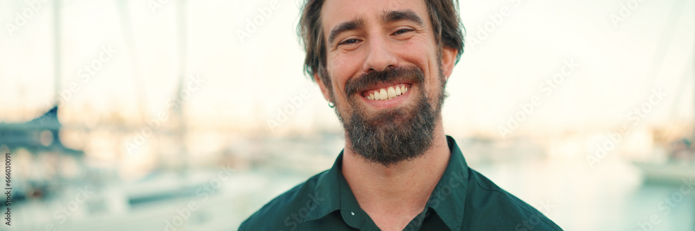 Obraz premium Close-up portrait of a smiling man with a beard on the embankment, on a yacht background. Frontal closeup of happy young hipster male looking at camera