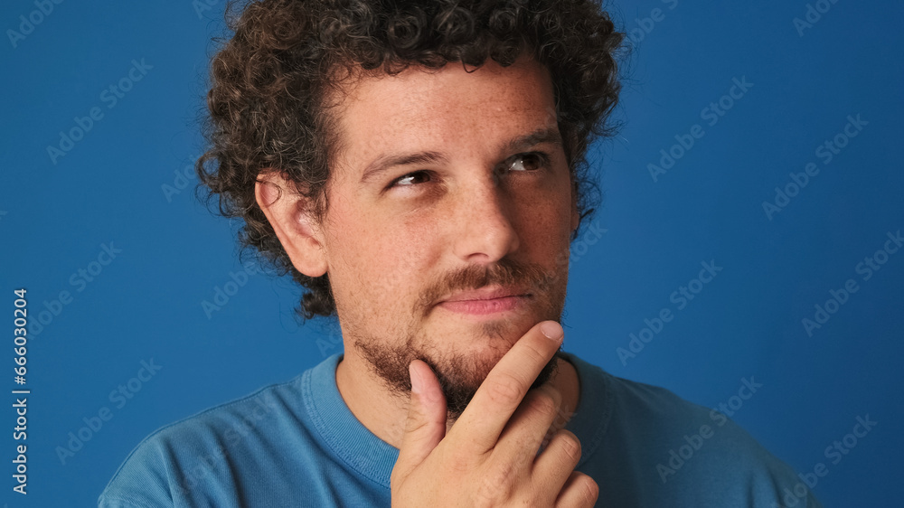 © Andrii Nekrasov - Close up, guy with curly hair dressed in blue t-shirt is thinking over difficult choice, imagining plan in his mind, having an idea, isolated on blue background in the studio