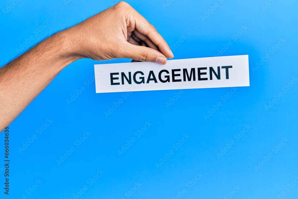 Hand of caucasian man holding paper with engagement word over isolated blue background Hand of caucasian man holding paper with engagement word over isolated blue background