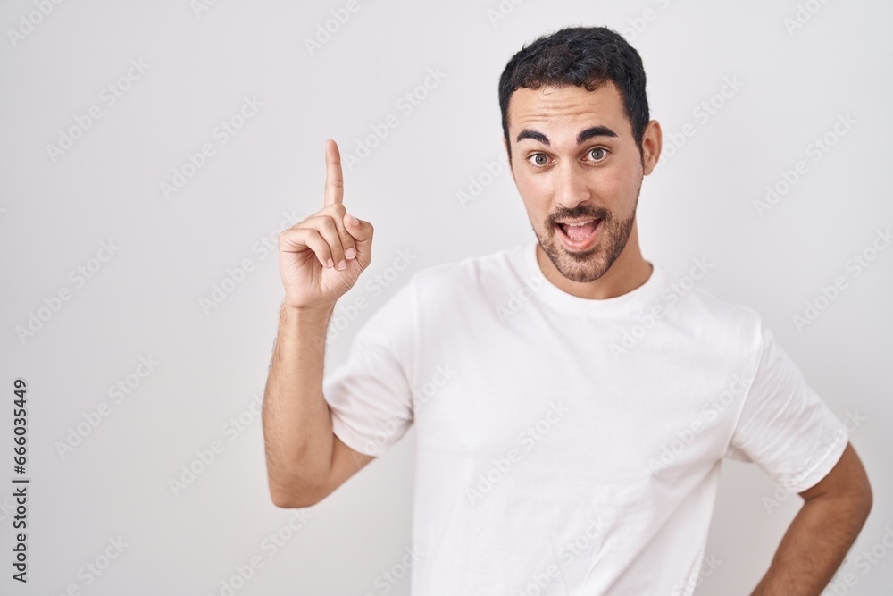 Handsome hispanic man standing over white background pointing finger up with successful idea. exited and happy. number one.