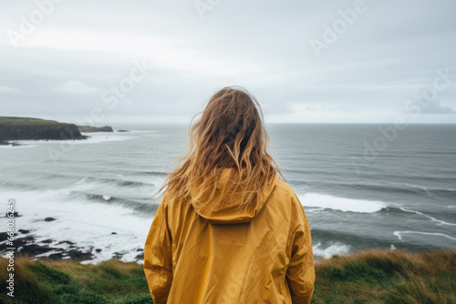 Young girl wearing raincoat standing on the edge of a cliff with huge waves rolling ashore. Rough Irish weather. Beautiful nature of Ireland.