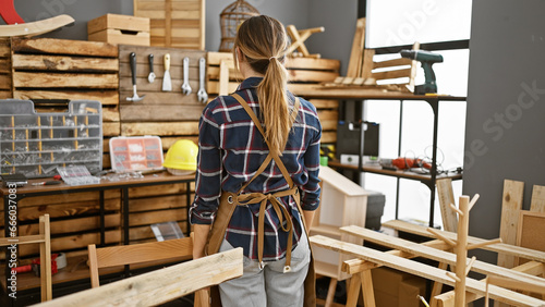 Entrancing back view of a young blonde carpenter  standing backwards  wearing her apron amidst a buzzing carpentry workshop