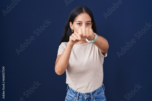 Young hispanic woman standing over blue background punching fist to fight, aggressive and angry attack, threat and violence