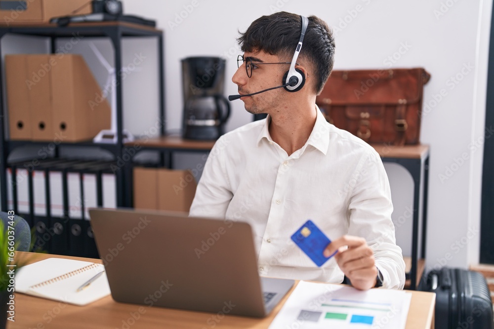 Young hispanic man working using computer laptop holding credit card looking to side, relax profile pose with natural face and confident smile.