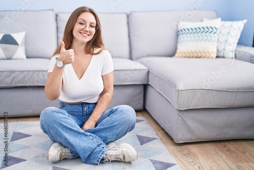 Young caucasian woman sitting on the floor at the living room doing happy thumbs up gesture with hand. approving expression looking at the camera showing success.