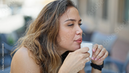 Young beautiful hispanic woman sitting on table holding cup coffee at coffee shop terrace