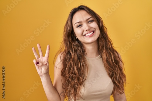 Young hispanic woman standing over yellow background showing and pointing up with fingers number three while smiling confident and happy.