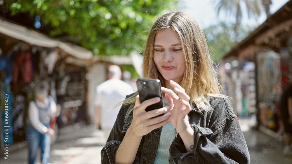 Young blonde woman using smartphone smiling at street market