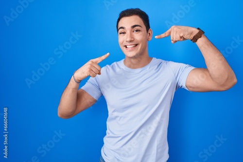 Young hispanic man standing over blue background smiling cheerful showing and pointing with fingers teeth and mouth. dental health concept.