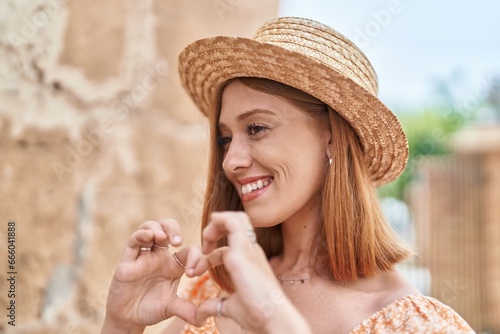 Young redhead woman tourist wearing summer hat doing heart gesture with hands at street
