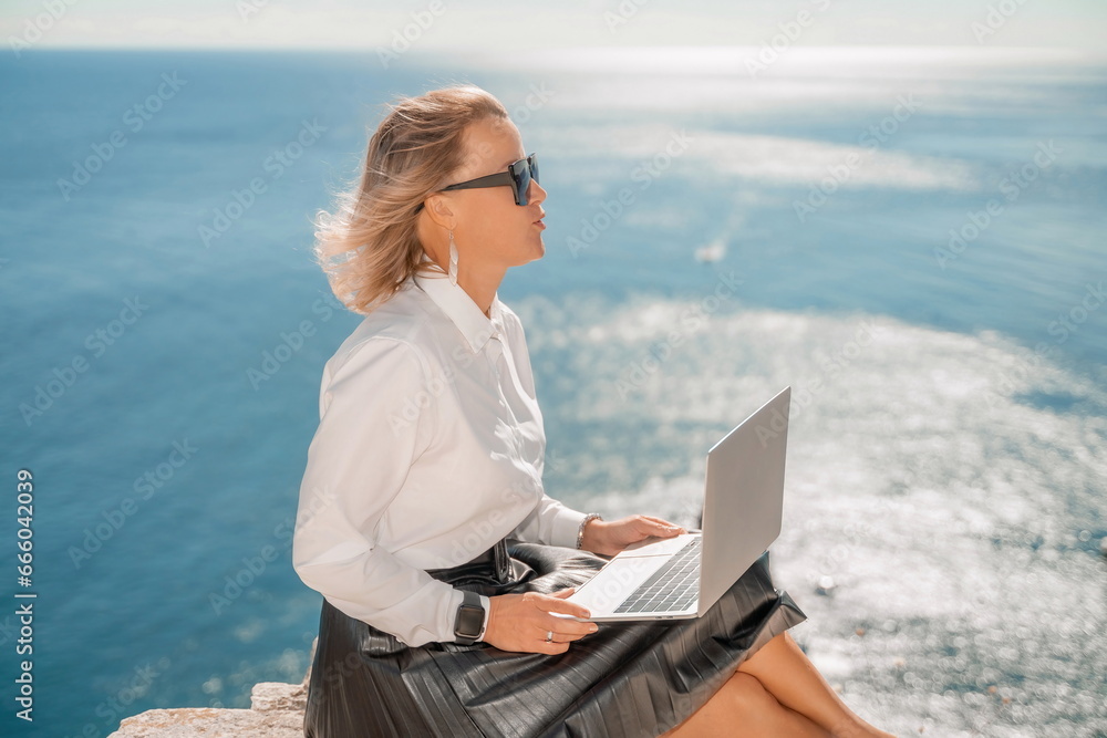 Business woman on nature in white shirt and black skirt. She works with an iPad in the open air with a beautiful view of the sea. The concept of remote work.