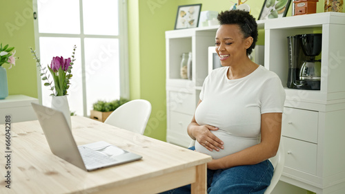 Young pregnant woman having video call sitting on table at dinning room