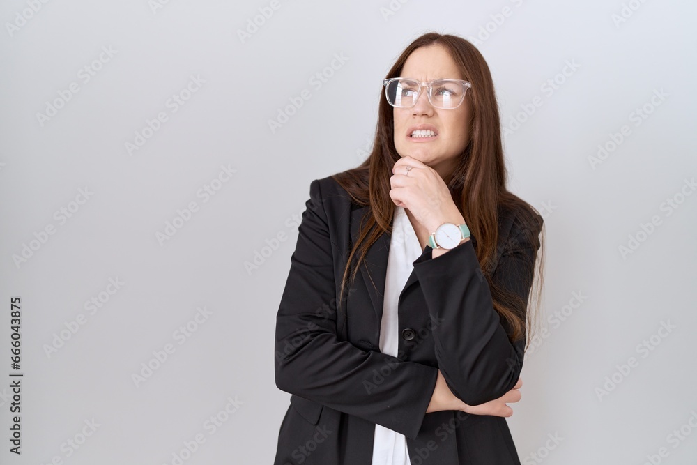 Beautiful brunette woman wearing business jacket and glasses thinking worried about a question, concerned and nervous with hand on chin Beautiful brunette woman wearing business jacket and glasses thinking worried about a question, concerned and nervous with hand on chin