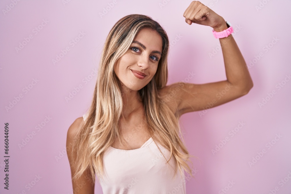 Young blonde woman standing over pink background strong person showing arm muscle, confident and proud of power Young blonde woman standing over pink background strong person showing arm muscle, confident and proud of power