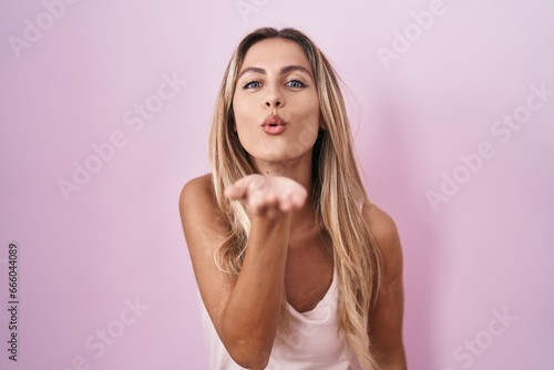 Young blonde woman standing over pink background looking at the camera blowing a kiss with hand on air being lovely and sexy. love expression.