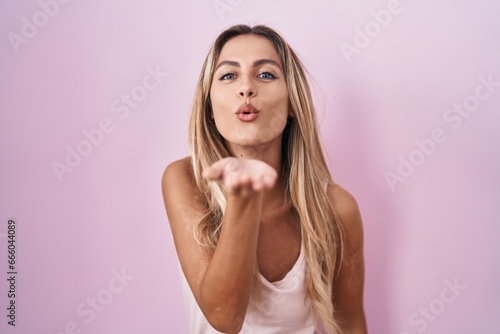Obraz na plátně Young blonde woman standing over pink background looking at the camera blowing a kiss with hand on air being lovely and sexy