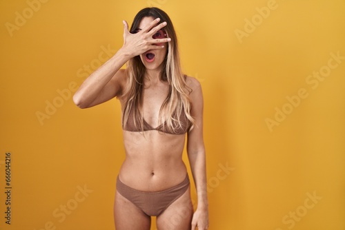 Young hispanic woman wearing bikini over yellow background peeking in shock covering face and eyes with hand, looking through fingers with embarrassed expression. © Krakenimages.com