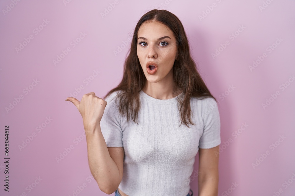 Young hispanic girl standing over pink background surprised pointing with hand finger to the side, open mouth amazed expression. Young hispanic girl standing over pink background surprised pointing with hand finger to the side, open mouth amazed expression.