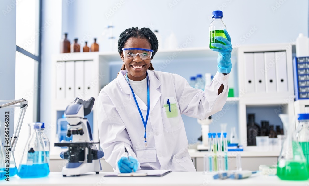 Fototapeta premium African american woman scientist holding bottle writing on document at laboratory
