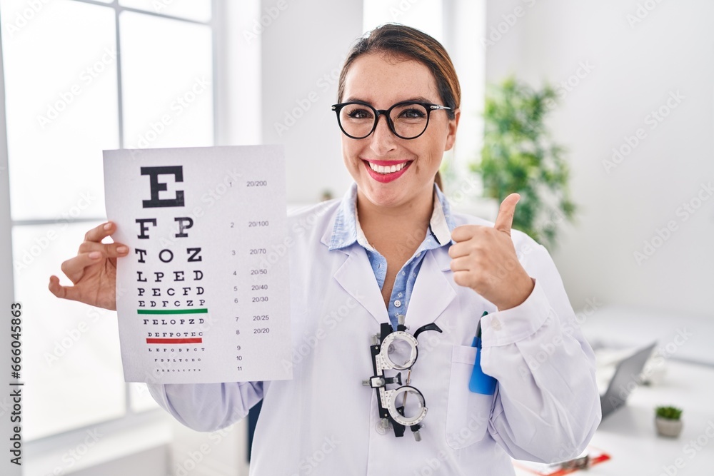 Young hispanic optician woman holding medical exam smiling happy and positive, thumb up doing excellent and approval sign Young hispanic optician woman holding medical exam smiling happy and positive, thumb up doing excellent and approval sign