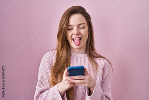 Young caucasian woman using smartphone typing message sticking tongue out happy with funny expression.