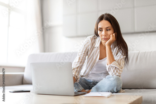 Upset Woman Looking At Laptop Computer While Sitting On Couch At Home © Prostock-studio