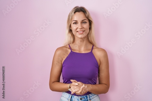 Young blonde woman standing over pink background with hands together and crossed fingers smiling relaxed and cheerful. success and optimistic