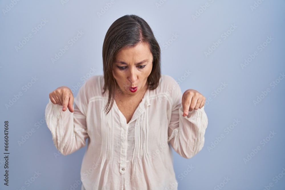 Middle age hispanic woman standing over blue background pointing down with fingers showing advertisement, surprised face and open mouth Middle age hispanic woman standing over blue background pointing down with fingers showing advertisement, surprised face and open mouth