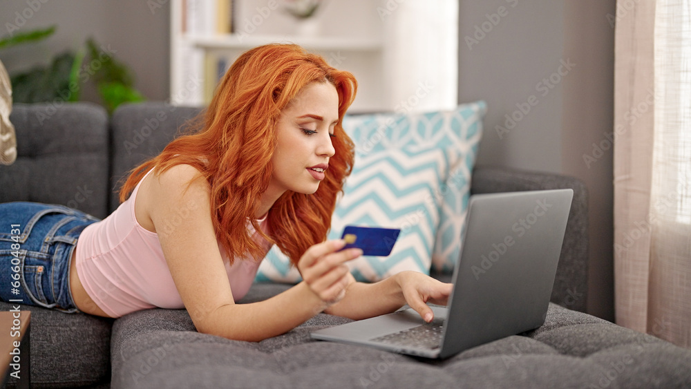 Young redhead woman shopping with laptop and credit card lying on sofa at home Young redhead woman shopping with laptop and credit card lying on sofa at home