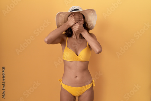 Young hispanic woman wearing bikini and summer hat covering eyes and mouth with hands  surprised and shocked. hiding emotion