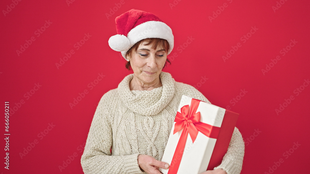 Mature hispanic woman smiling holding present wearing christmas hat over isolated red background