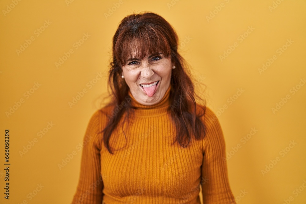 Middle age hispanic woman standing over yellow background sticking tongue out happy with funny expression. emotion concept. Middle age hispanic woman standing over yellow background sticking tongue out happy with funny expression. emotion concept.