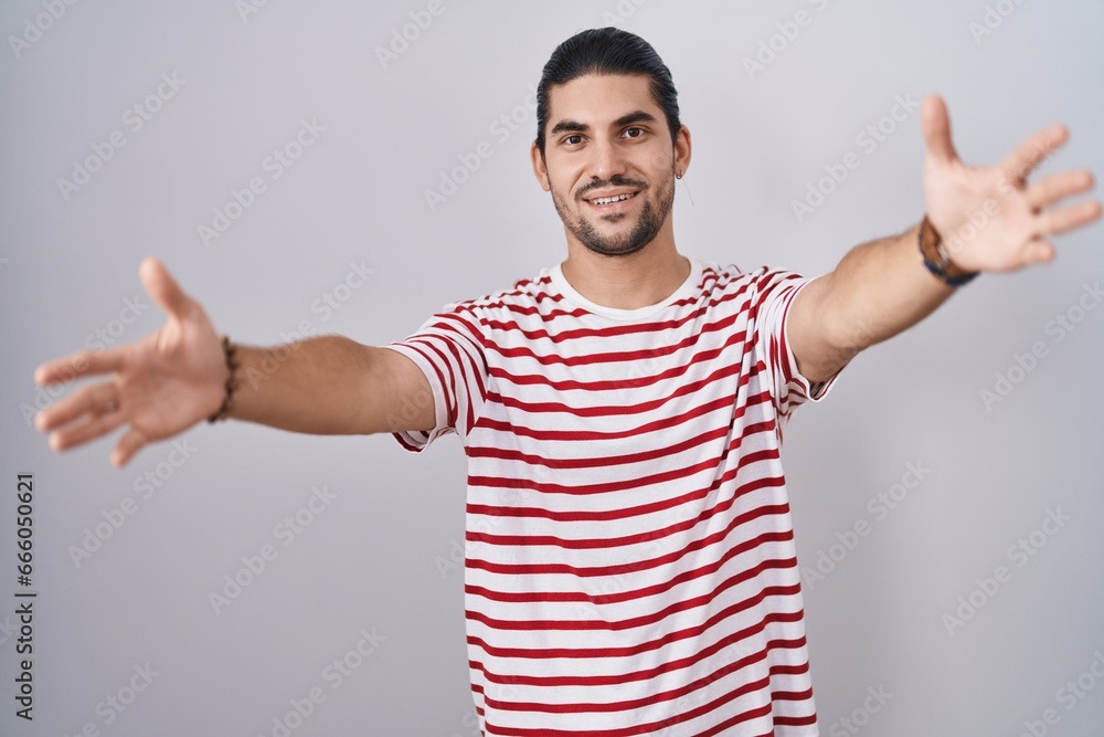Hispanic man with long hair standing over isolated background looking at the camera smiling with open arms for hug. cheerful expression embracing happiness. Hispanic man with long hair standing over isolated background looking at the camera smiling with open arms for hug. cheerful expression embracing happiness.