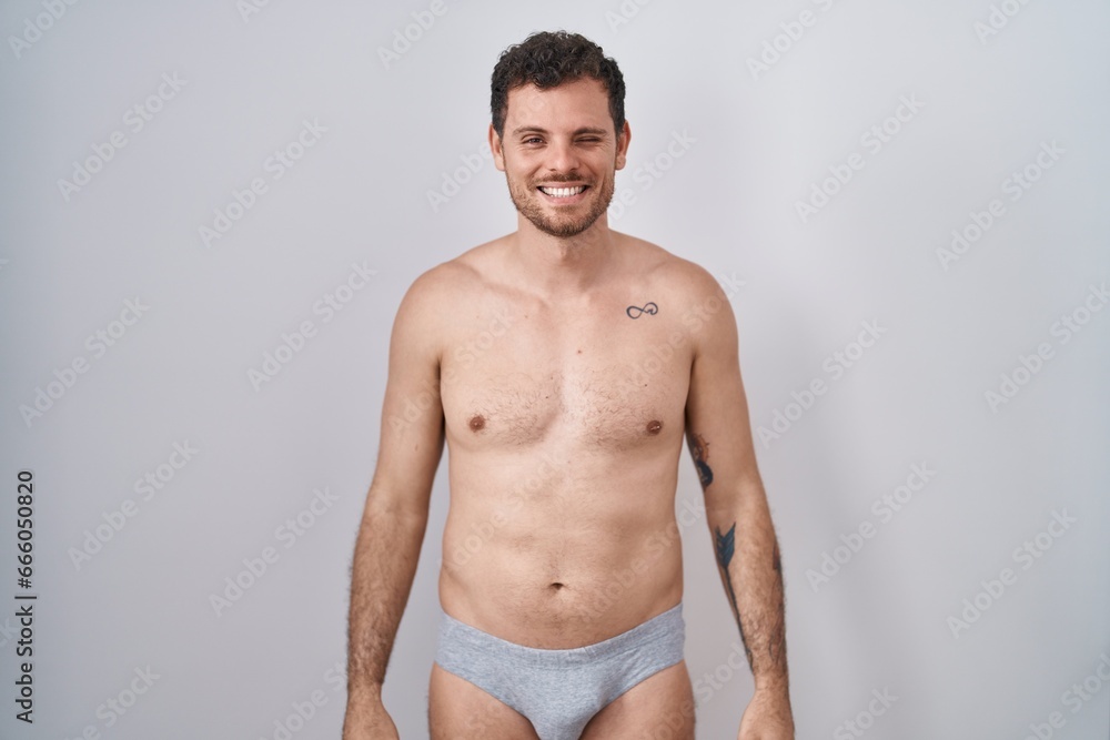 Fototapeta premium Young hispanic man standing shirtless wearing underware winking looking at the camera with sexy expression, cheerful and happy face.
