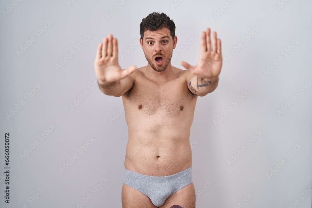 Young hispanic man standing shirtless wearing underware doing stop gesture with hands palms, angry and frustration expression