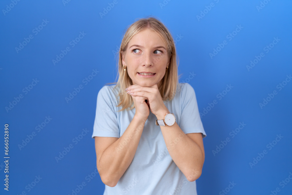 Young caucasian woman wearing casual blue t shirt laughing nervous and excited with hands on chin looking to the side Young caucasian woman wearing casual blue t shirt laughing nervous and excited with hands on chin looking to the side