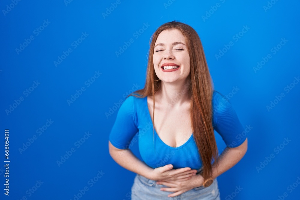 Redhead woman standing over blue background smiling and laughing hard out loud because funny crazy joke with hands on body. Redhead woman standing over blue background smiling and laughing hard out loud because funny crazy joke with hands on body.