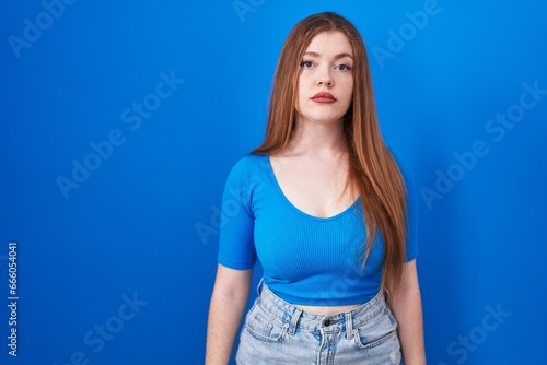 Redhead woman standing over blue background relaxed with serious expression on face. simple and natural looking at the camera.