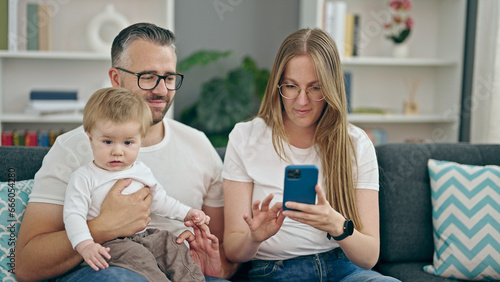 Family of mother, father and baby smiling using smartphone sitting on the sofa at home