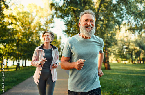 Running in park at morning time. Cheerful husband and wife competing together and jogging on fresh air. Active people wearing sport clothes doing cardio for good health and staying fit. photo