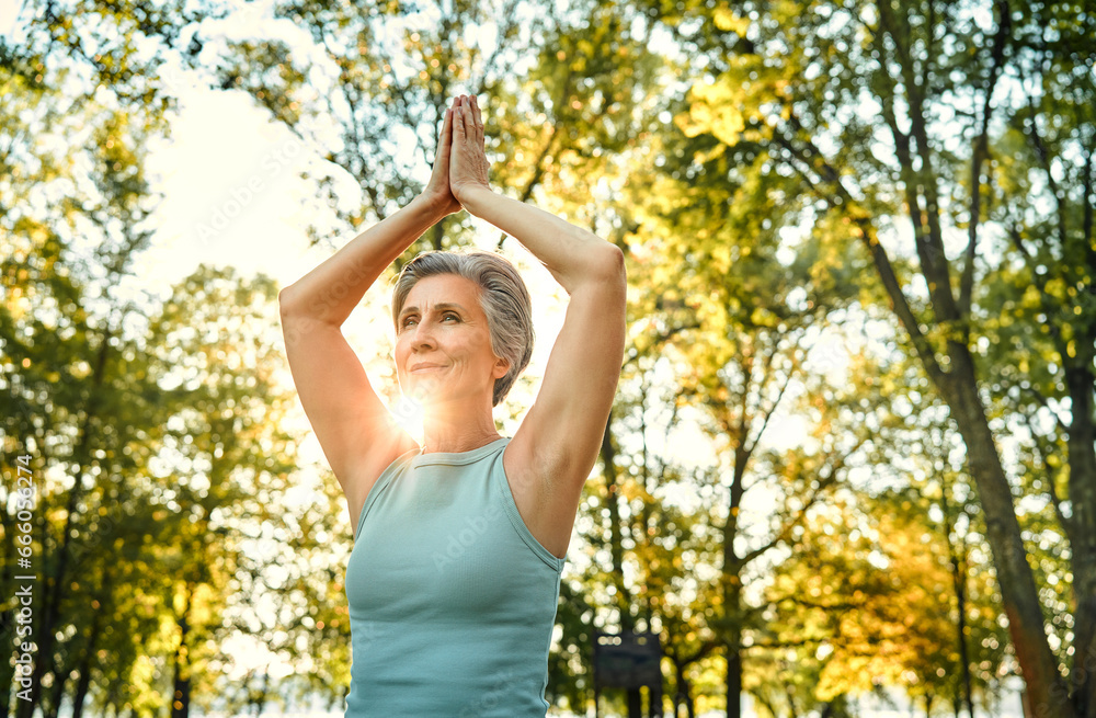 Yoga practice outdoors. Graceful aged lady keeping hands clasped above head and making regular workout on fresh air. Grey-haired woman wearing sport attire performing vrksasana tree pose. Yoga practice outdoors. Graceful aged lady keeping hands clasped above head and making regular workout on fresh air. Grey-haired woman wearing sport attire performing vrksasana tree pose.