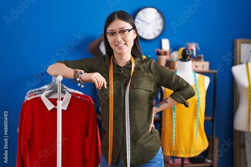 Chinese woman tailor smiling confident leaning on clothes rack at sewing studio