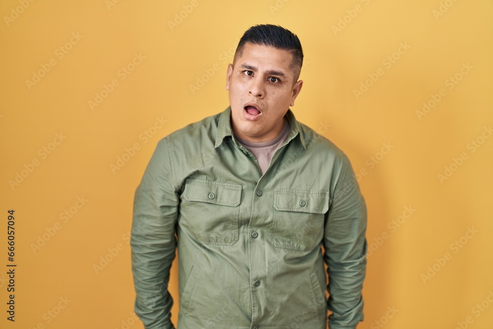 Fototapeta premium Hispanic young man standing over yellow background in shock face, looking skeptical and sarcastic, surprised with open mouth